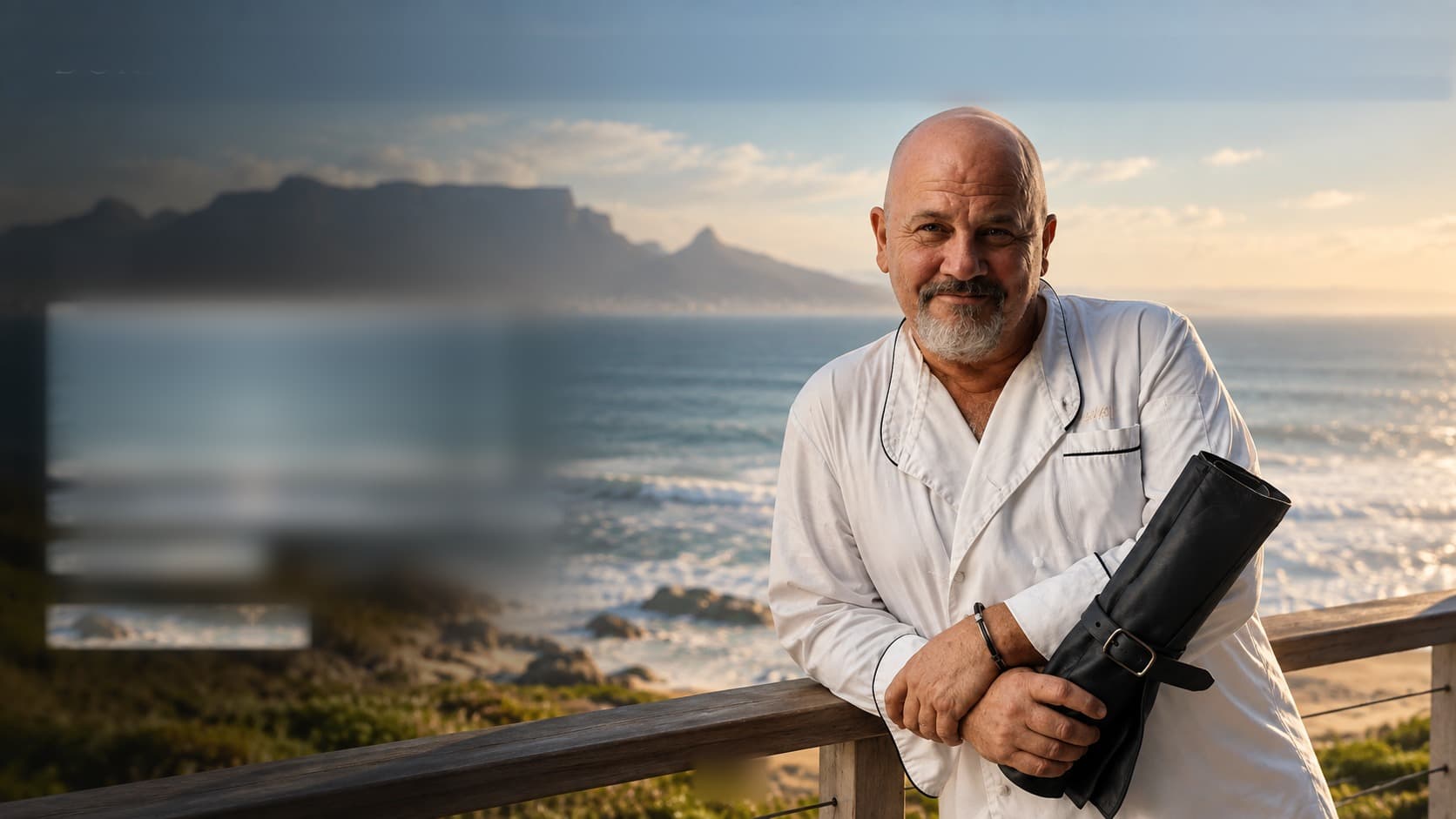 Chef Paul Dukai on the Cape coast at golden hour, holding his knife roll, Table Mountain across the bay