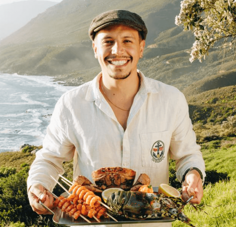 Flavio Mariano smiling in a flat cap and linen shirt, holding a tray of grilled prawn skewers, crab and lobster above a Cape coastline