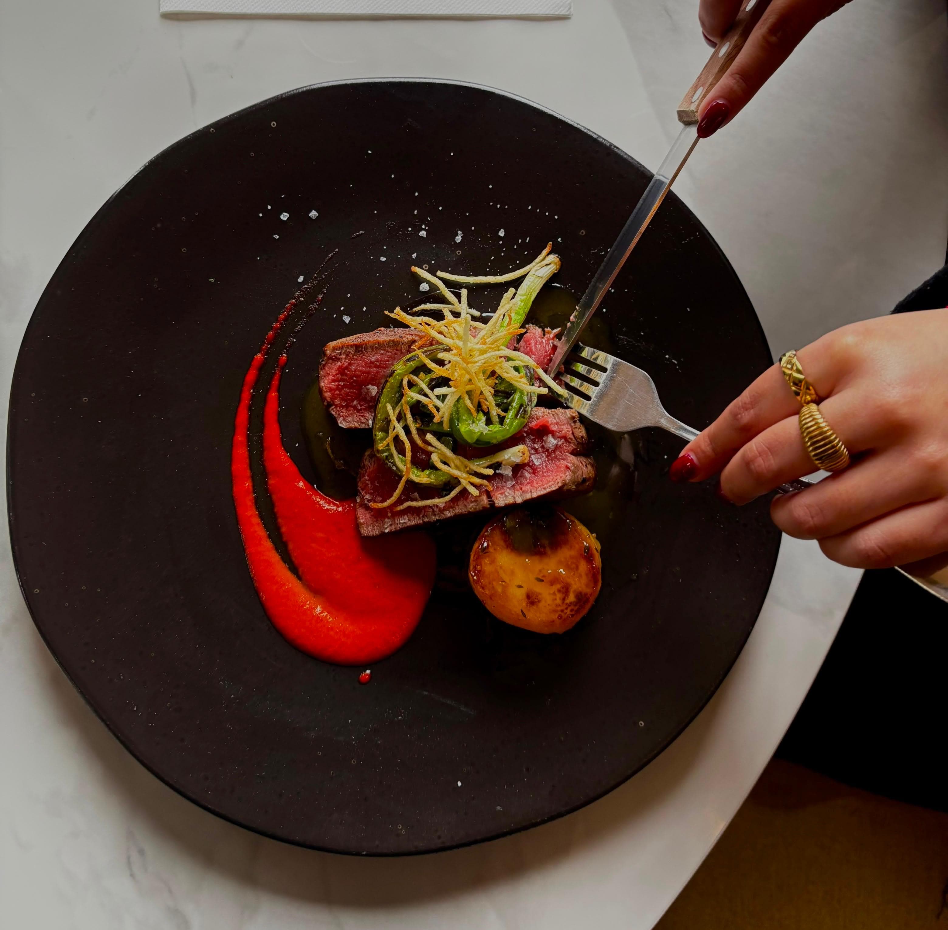 Sliced beef fillet, cooked medium rare, with a sweep of piquillo pepper purée, a glazed potato fondant, charred spring onion and crisp potato threads on a dark plate