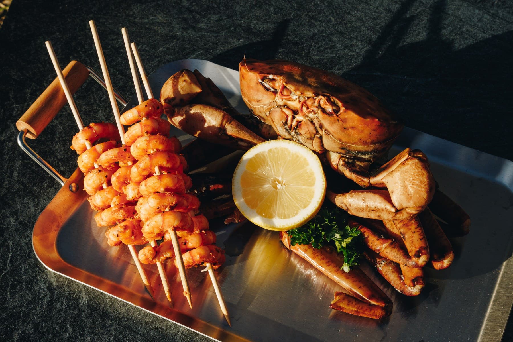 Grilled prawn skewers beside a cooked crab and lemon on a steel tray